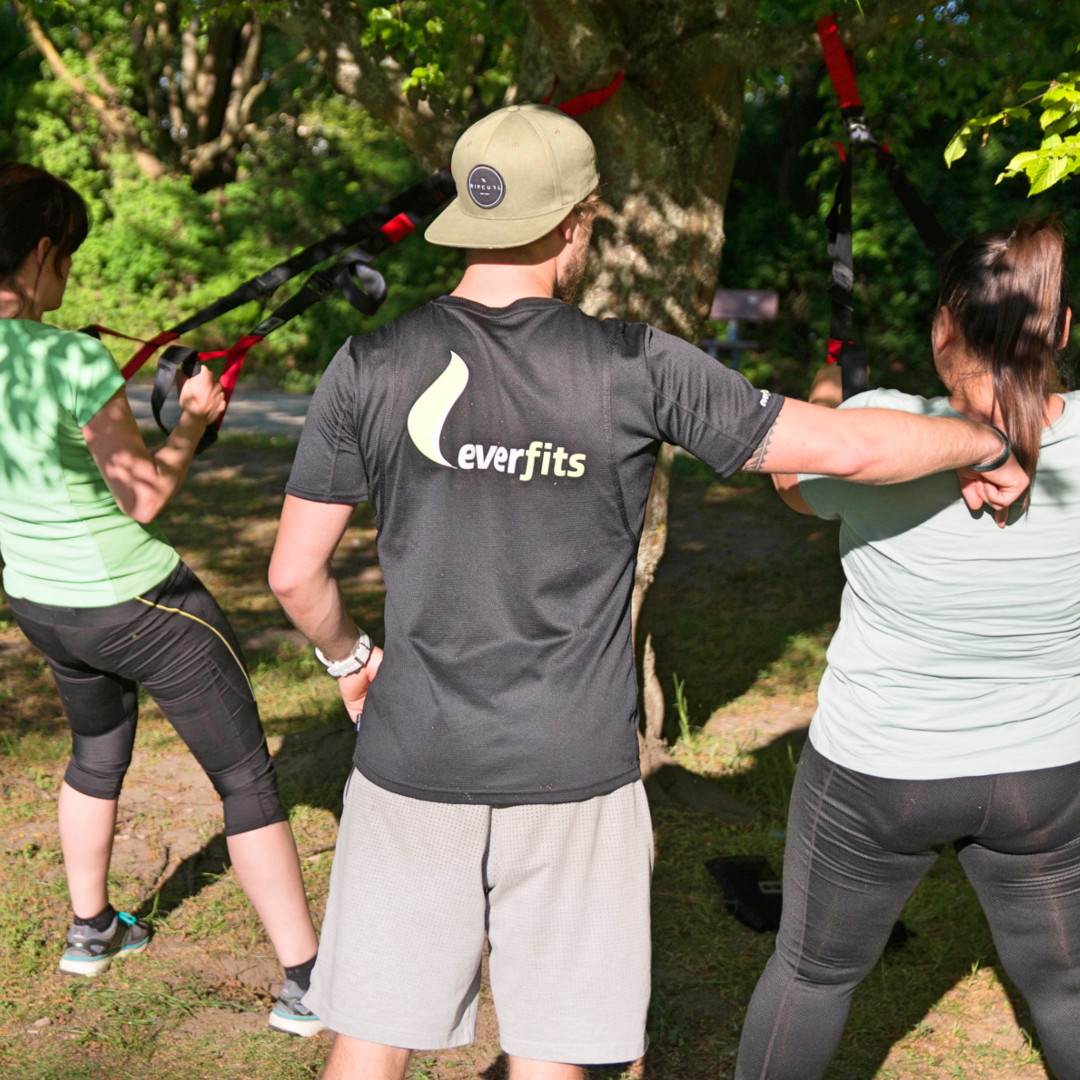 Fitness Trainer korrigiert beim Outdoor Training die Ausführung der Übung Rudern mit dem Slingtrainer